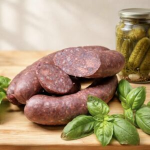 Wiejska kaszanka gryczana prezentowana na jasnej, drewnianej desce. Dwa całe pęta o gładkiej strukturze są ułożone obok rzędu grubych, skośnie uciętych plastrów, które pokazują bogate wnętrze z kawałkami mięsa. W tle stoi słoik tradycyjnych ogórków kiszonych z koperkiem, a całość uzupełniają świeże liście bazylii. Estetyczne ujęcie produktowe idealne do delikatesów online.