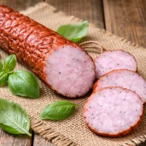 Zbliżenie na apetyczne plastry i przekrój wędzonej szynki lub kiełbasy z widocznymi kawałkami mięsa i przypraw, ułożonych na lnianej ściereczce w otoczeniu świeżych liści bazylii.