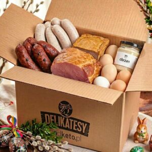 Otwarte kartonowe pudełko z logo "Delikatesy podketo.pl" wypełnione wielkanocnymi produktami: białą kiełbasą, kiełbasą myśliwską, plastrami wędliny, kawałkiem pasztetu, słoiczkiem chrzanu oraz jajkami. Pudełko stoi na obrusie w scenerii wielkanocnej z pisankami, baziami i figurką zajączka.